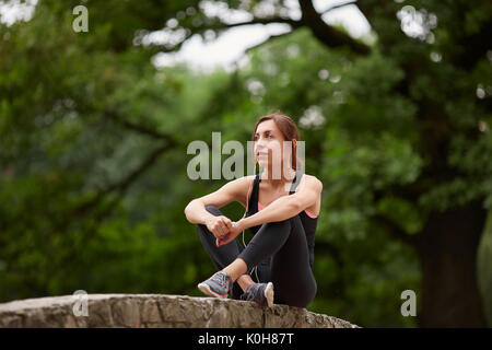 Young content woman in sportswear sitting on stone fence in park and listening to music in headphones. Fitness woman in city park. Caucasian fitness m Stock Photo