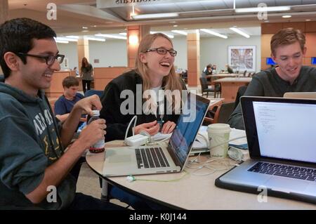 Three students, two males on either side of a female, seated on a table in the second level, M Level, of the Milton S. Eisenhower Library, surrounded by study materials and laptops, other students depicted in the background, all with smiling facial expressions, 2016. Courtesy Eric Chen. Stock Photo