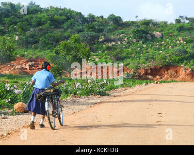 A girl child going to school on a sunny day with her bicycle. A school bag of the child can also be seen in the picture. Stock Photo