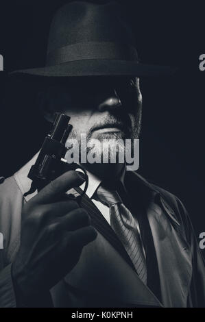 1950s style detective posing in the dark, he is holding a weapon and wearing a fedora hat, noir film character Stock Photo