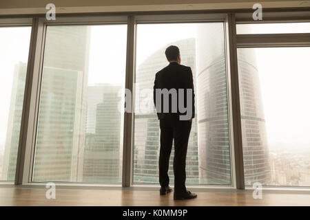 Businessman standing near full-length window looking at city, ba Stock Photo