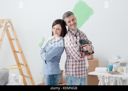 Smiling loving couple doing home renovations, the woman is holding a paint roller and the man is using a drill Stock Photo