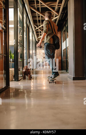 Full length rear view shot of young man skateboarding in modern office. Casual businessman skating through his startup office. Stock Photo