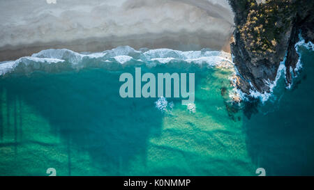 Drone shot of a rocky headland at sunset with small breaking waves in emerald blue water. Stock Photo