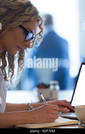Close up of young woman writing on book while using laptop at table in cafe Stock Photo