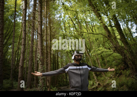 Man wearing vr glasses while standing with arms outstretched amidst trees in forest Stock Photo