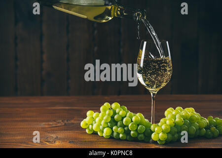 Pouring white wine from the bottle into a glass with a bunch of green grapes against wooden background with free space Stock Photo