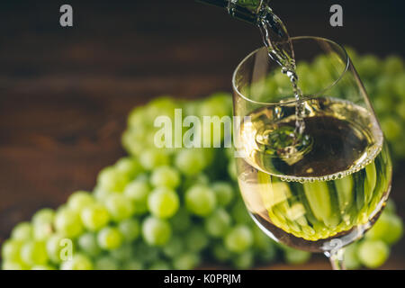 Pouring white wine into a glass with a bunch of green grapes against wooden background Stock Photo