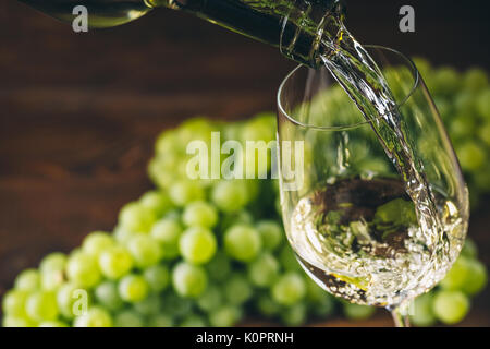 Pouring white wine into a glass with a bunch of green grapes against wooden background Stock Photo