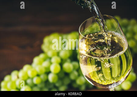 Pouring white wine into a glass with a bunch of green grapes against wooden background Stock Photo