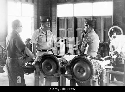 African-american lieutenant standing with two other American servicemen during the Vietnam War, in a heavy vehicle machine shop, one man smoking a cigarette, the other man resting his hand on a part for a truck, the lieutenant gesturing and in mid speech, 1969. Stock Photo