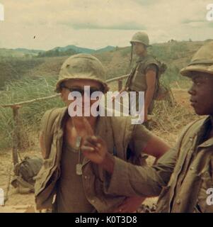 Marine in combat vest and helmet, wearing sunglasses and dog tag, standing in a field during the Vietnam war, an African-American fellow Marine pointing to him as a third Marine stands in the background, 1965. Stock Photo