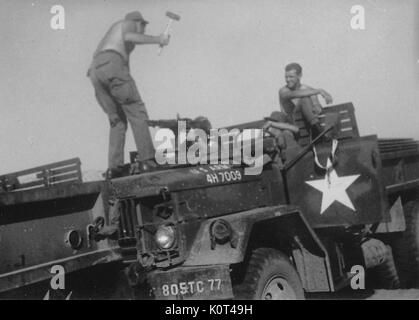 A photograph of four United States Army serviceman amusing each other on a parked M35 military truck, two of the soldiers watch in amusement while a soldier with a hammer stands on the hood of the truck in front another soldier who has a hand on a mounted M60 machine gun, Vietnam, 1967. Stock Photo