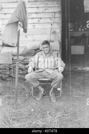 A United States Army serviceman relaxing in a lawn chair, he is smoking a cigarette near the entrance to a wooden building where equipment is visible spread out on a table, Thailand, 1967. Stock Photo