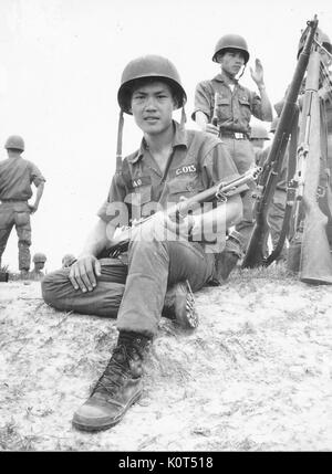 A photograph of an Army of the Republic of Vietnam soldier holding his combat rifle, other soldiers are visible in the background as well as a several guns leaned together in an upright position, Vietnam, 1968. Stock Photo