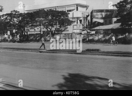 A photograph of two men traveling along a paved road that runs through a residential area, one is walking while the other is riding a bicycle, multi-story buildings are seen on the opposite side of the street, Vietnam, 1967. Stock Photo