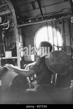 A United States Army serviceman seated near a large window on an airplane, he has his gear between his legs and a helmet hanging from his chair, Vietnam, 1967. Stock Photo