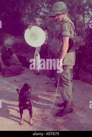 A group of United States Servicemen in uniform are gathered together in a small group, one soldier has a dog on a leash while another soldier who is a member of the 1st Infantry Division Band leans on a tuba, other soldiers can be seen in the background, Vietnam, 1968. Stock Photo