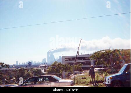 During the terrorist attacks of September 11, 2001, the Twin Towers of the World Trade Center in Manhattan are seen from Jersey City, New Jersey (3 miles West of Ground Zero), where a mature woman stands behind a fence and looks at a plume of black smoke rising from the buildings, September 11, 2001. Stock Photo