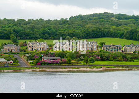Waterfront buildings of town of Rothesay at foot of hill with green fields and woods and beside calm water of ocean at island of Bute, Scotland Stock Photo