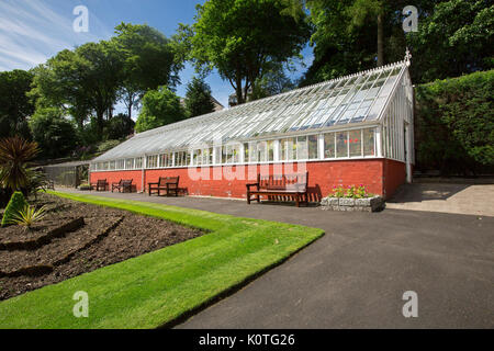 Greenhouse at Ardencraig gardens on Bute, Scotland, with red painted wall lower section, lawns in foreground, and trees in background under blue sky Stock Photo