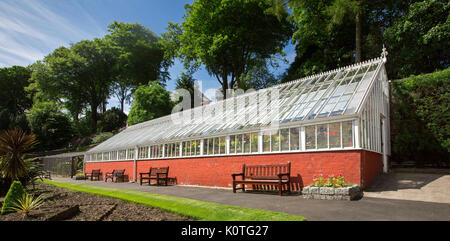 Greenhouse at Ardencraig gardens on Bute, Scotland, with red painted wall lower section, lawns in foreground, and trees in background under blue sky Stock Photo