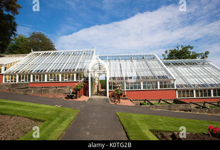 Ornate greenhouse complex at Ardencraig gardens on Bute, Scotland, with red painted wall lower section &lawns in foreground under blue sky Stock Photo