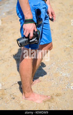 Barefoot photographer in blue shorts on a sandy beach. DSLR camera in hand in sea beach background Stock Photo