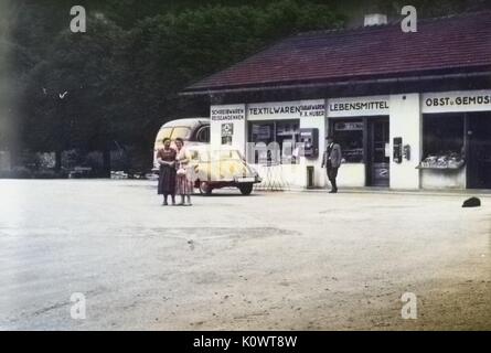 Two women and a man outside a roadside shop, a car parked in front, Germany, 1946. Note: Image has been digitally colorized using a modern process. Colors may not be period-accurate. Stock Photo
