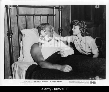 A movie still scene from 'The Tougher They Come' (1950 Columbia Pictures film), showing an injured man with bandages on the left side of his head and his left arm in a cast lying in bed while a young woman puts her right hand on his forehead to check his temperature, 1950. Stock Photo