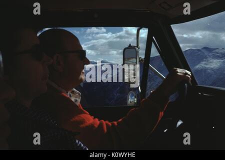 Mature tourists driving in a pickup truck through the mountains, a man and a woman, viewed from within the cab of the truck, the man with his hands on the steering wheel, in silhouette, with a scene of snow capped mountain peaks visible through the window of the truck, 1975. Photo credit Smith Collection/Gado/Getty Images. Stock Photo