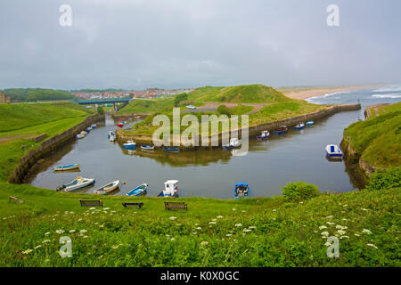 Boats moored in sheltered harbour at Seaton Sluice near coastal village of Hartley, with road bridge, town, & sandy beach nearby, England