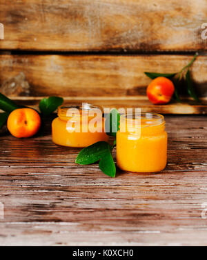 Apricot jam in small glass jars with fruits and leaves of apricot and twigs on rustic table, selective focus Stock Photo