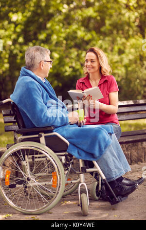 young daughter reading book outdoor her disabled father in wheelchair Stock Photo