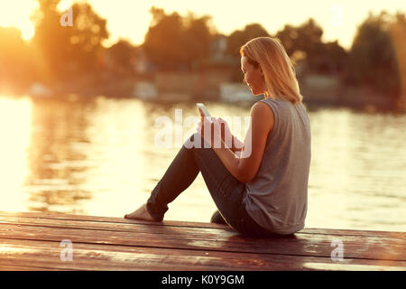 Girl with cellphone sitting on dock near river on sunset Stock Photo