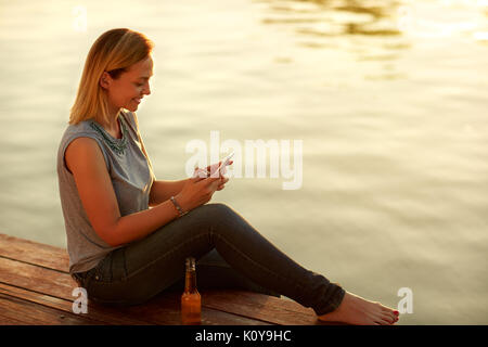 Young smile woman sitting on dock and looking at celphone Stock Photo