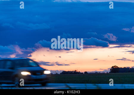 Speeding motion blur oncoming car with glowing lights on the highway after sunset. Shining the spotlight cars. Blue and orange bright sky at dusk. Abs Stock Photo
