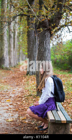 Pretty beautiful blonde child schoolgirl back to school sitting on bench in tree alley in nature with backpack, glasses, white shirt and purple skirt  Stock Photo