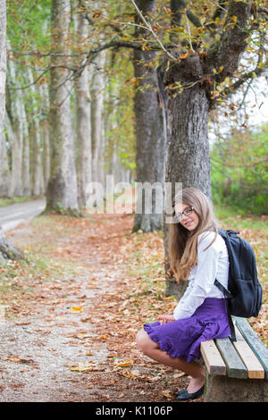 Pretty beautiful blonde child schoolgirl smiling back to school sitting on bench in tree alley in nature with backpack, glasses, white shirt and purpl Stock Photo