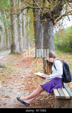 Pretty little blonde schoolgirl with backpack and long hair sitting on the bench in the alley park and reading a book getting ready to go back to scho Stock Photo