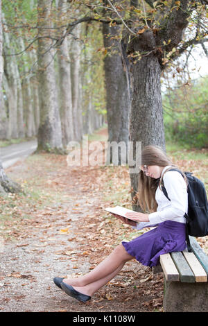 Pretty little blonde schoolgirl with backpack and long hair sitting on the bench in the alley park and reading a book getting ready to go back to scho Stock Photo