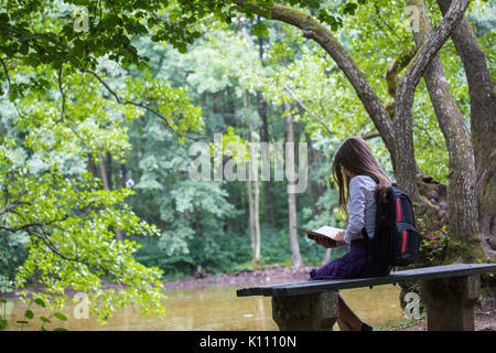 Pretty little blonde schoolgirl with backpack sitting on the bench in the nature park near lake and reading a book getting ready to go back to school  Stock Photo