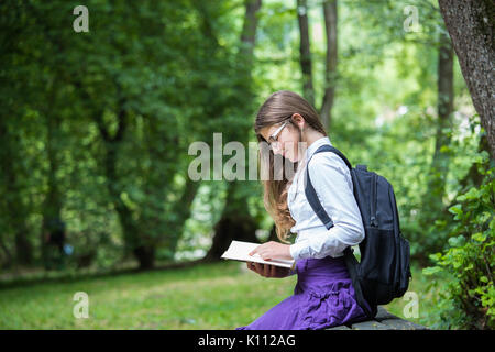 Pretty little blonde schoolgirl with backpack and long hair sitting on the bench in the nature park and reading a book ready to go back to school in t Stock Photo