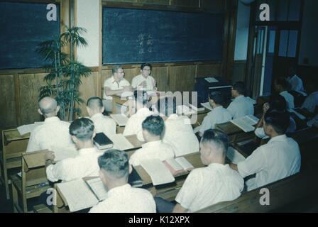 Japanese bible study class, large group of Japanese men in white shirts sitting in a rows in a classroom reading prayer books, a Caucasian missionary and Japanese man sitting at the front of the classroom and gesturing, Japanese characters visible on blackboard, at Wesleyan Methodist Mission, Japan, 1952. Stock Photo