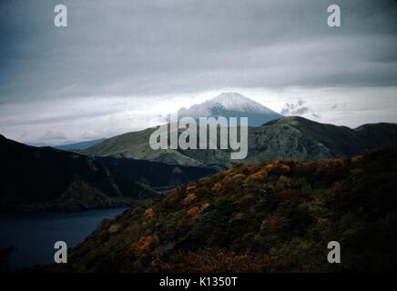 Mount Fuji and Lake Hakone, luminescent orange foliage lit by the sun in the foreground, snow-capped peak of the mountain visible in the background, with low cloud cover on an overcast day, Japan, November 8, 1951. Stock Photo