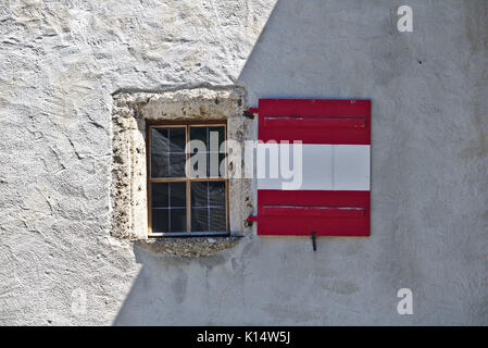 Rough plastered castle wall with window and wooden shutters colored like the Austrian flag, partly shaded and partly lit by sunlight Stock Photo