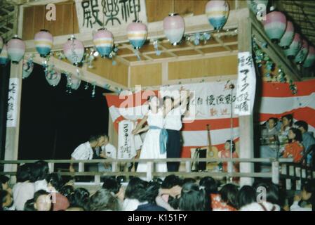Kabuki theater performance, a man and woman dancing together on stage, musicians visible in the background, crowd visible in the foreground watching the performance, hanging lanterns decorating the stage, Japan, 1951. Stock Photo