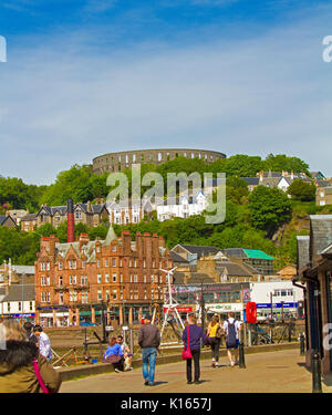 Scottish town of Oban with McCaig's tower on hilltop and people wandering beside harbour and waterfront buildings Stock Photo