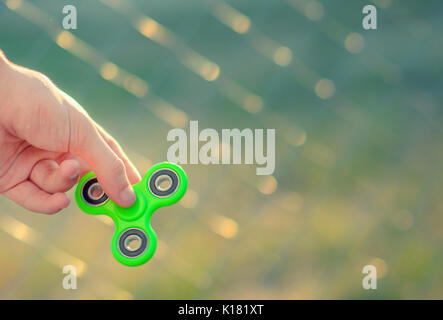 Young man hand holding antistress modern gadget fidget spinner. Teenager playing with green spinner outdoors on the rabitz grid bokeh. Shallow DOF. Stock Photo