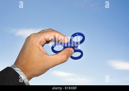 Male hand holding a fidget spinner. Blue spinner in the hand of a businessman in coat on the background of blue sky Stock Photo